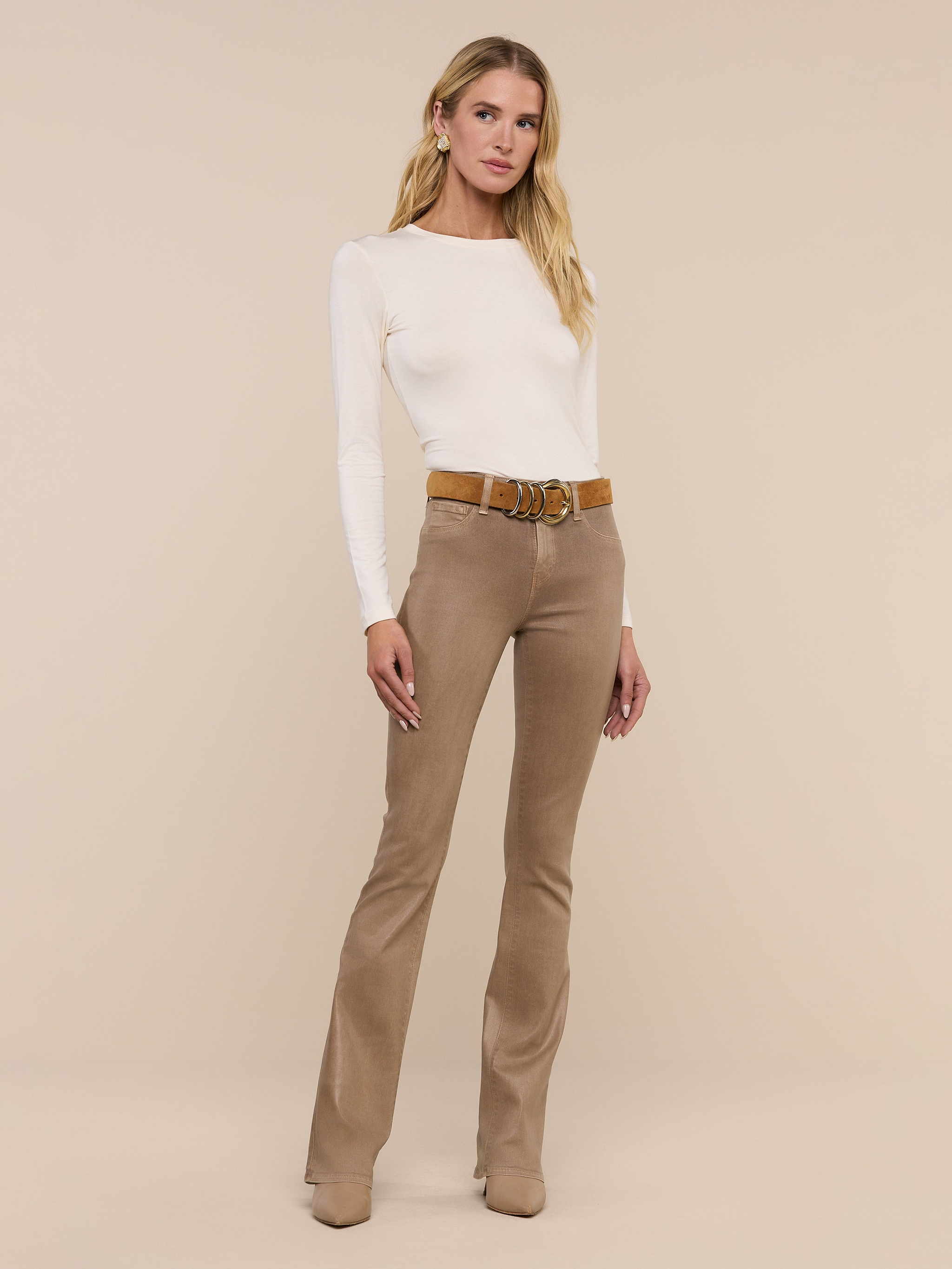 A woman with long blonde hair stands against a plain white background, wearing the Tess Long Sleeve Tee in a slim fit, paired with tan flared pants, a brown belt, and tan boots as she looks at the camera.
