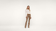 A woman stands in front of a plain white background, wearing a long-sleeve white top, tan pants, tan belt, and tan shoes. She has long blonde hair and is looking at the camera with a neutral expression.