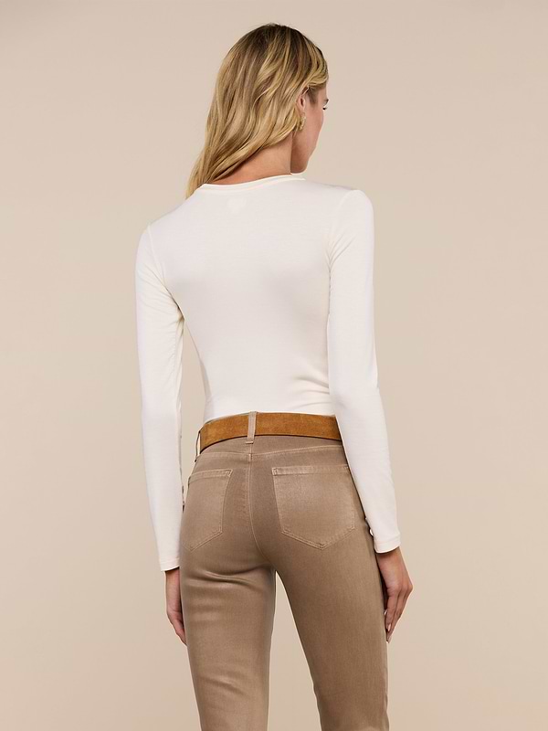 A person with long blonde hair is shown from the back, wearing the Tess Long Sleeve Tee in a slim fit cream shade and beige pants with a brown belt, standing against a plain light background.