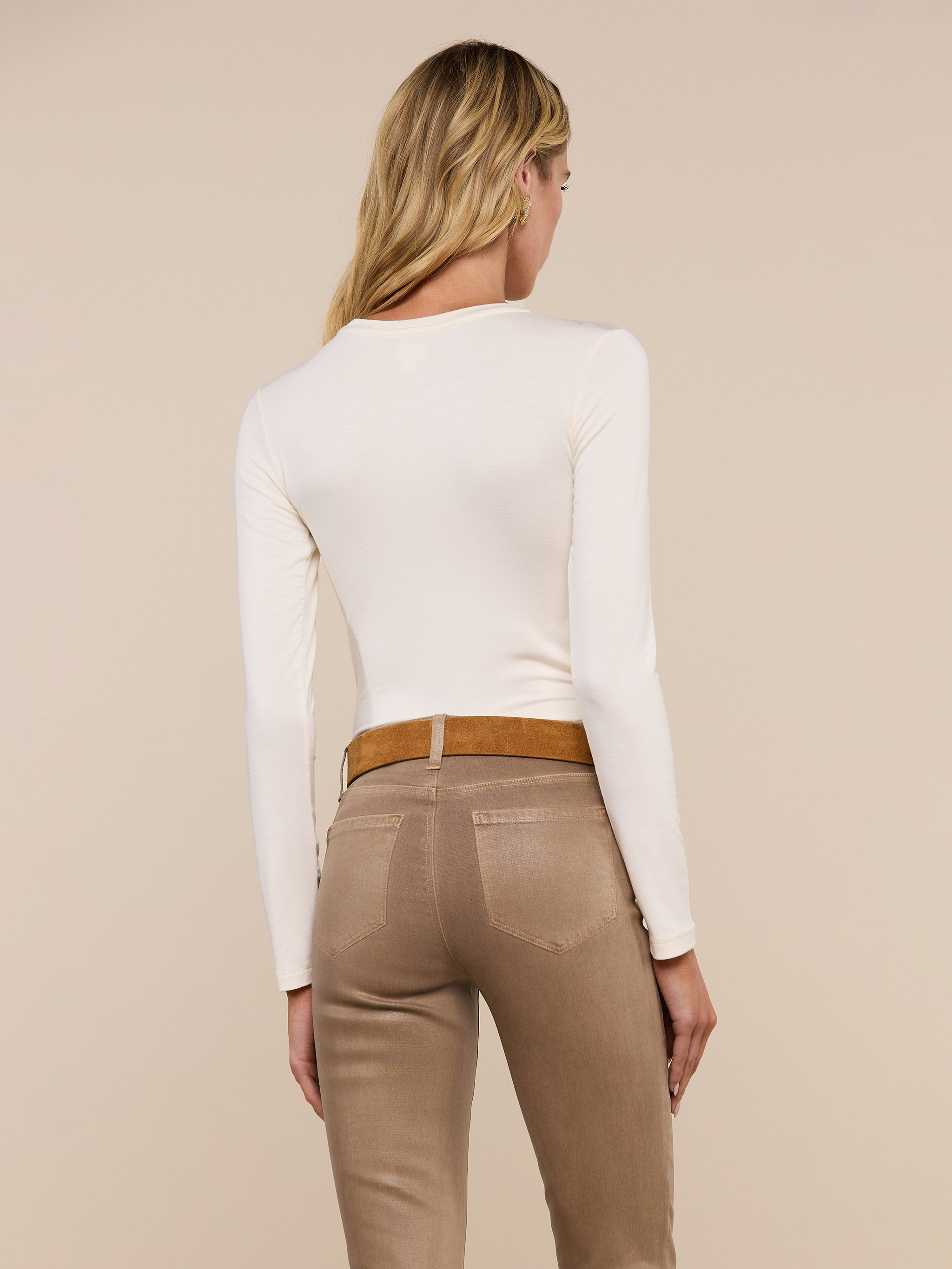 A person with long blonde hair is shown from the back, wearing the Tess Long Sleeve Tee in a slim fit cream shade and beige pants with a brown belt, standing against a plain light background.