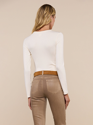 A woman with long blonde hair, wearing a slim fit Tess Long Sleeve Tee in white, tan jeans, and a brown belt, stands facing away from the camera against a beige background.