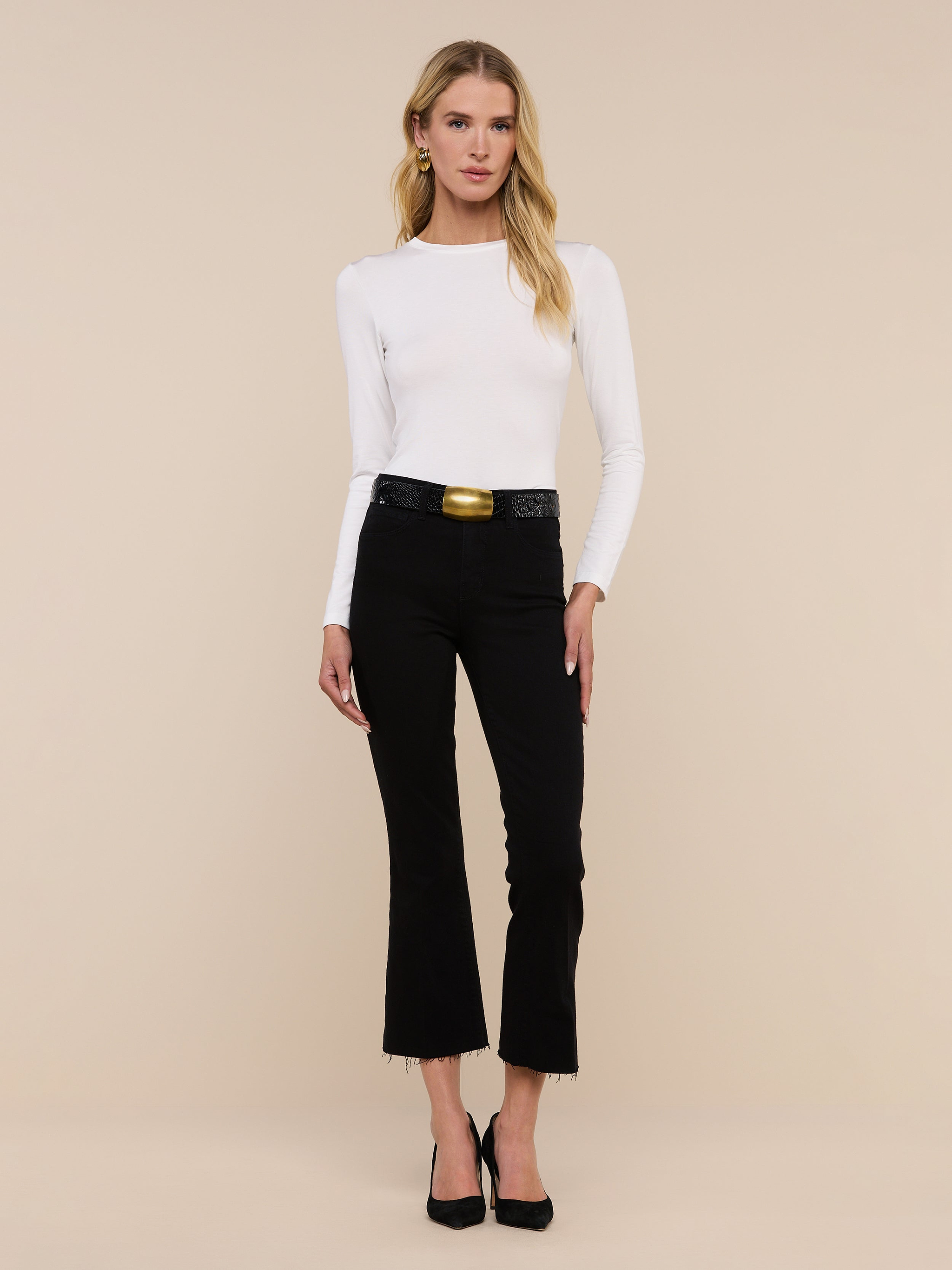 A woman stands facing forward against a plain white background, wearing the Tess Long Sleeve Tee—a slim-fit, microfiber jersey top in white—paired with black pants with a belt and black heels. The Tess Long Sleeve Tee offers a versatile layering option that adds a sleek touch to her outfit.