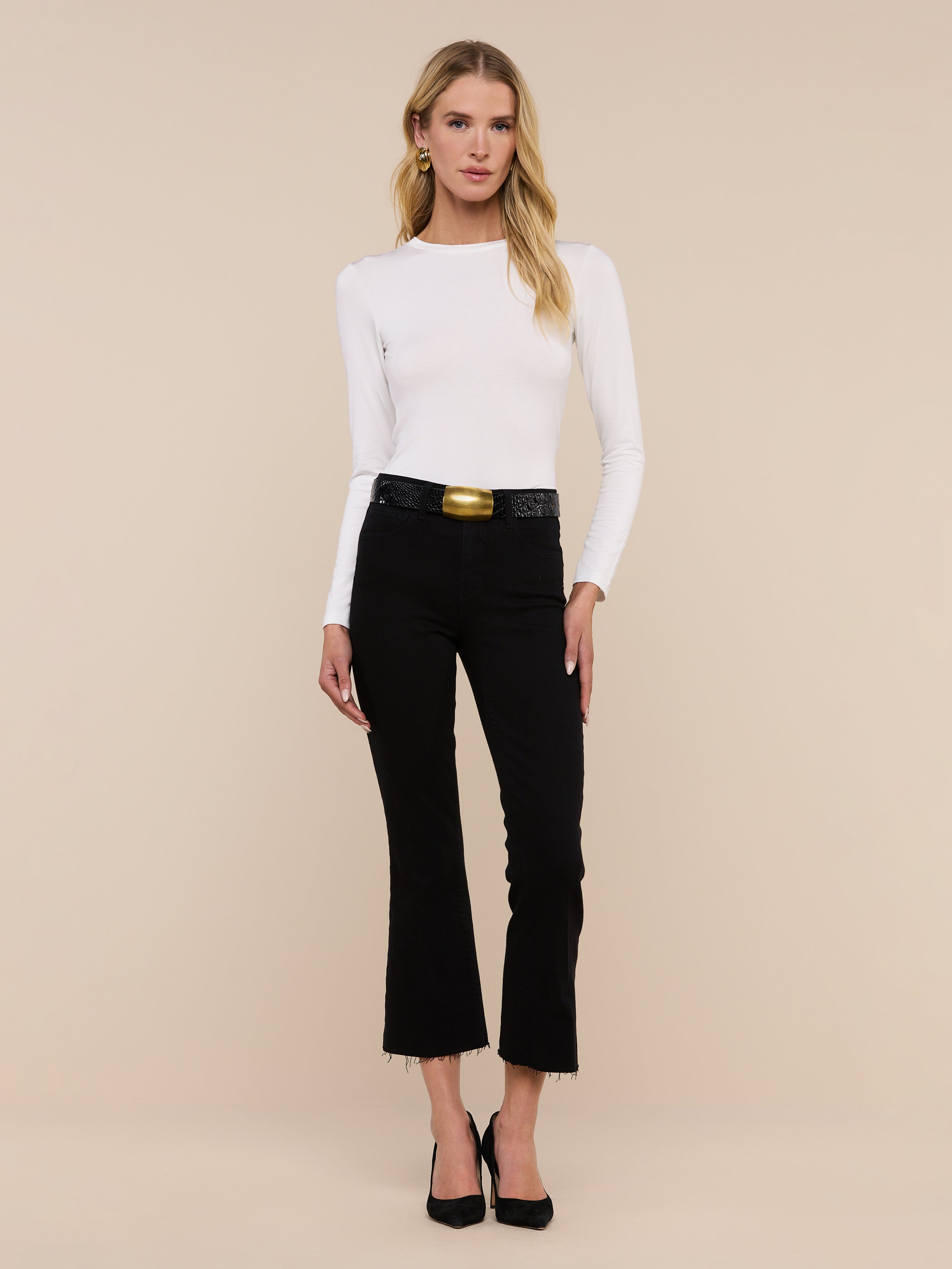 A woman stands facing forward against a plain white background, wearing the Tess Long Sleeve Tee—a slim-fit, microfiber jersey top in white—paired with black pants with a belt and black heels. The Tess Long Sleeve Tee offers a versatile layering option that adds a sleek touch to her outfit.