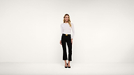 A woman stands in a minimal, white studio wearing a fitted white top, black pants with a belt, and black heels, facing forward with a neutral expression.