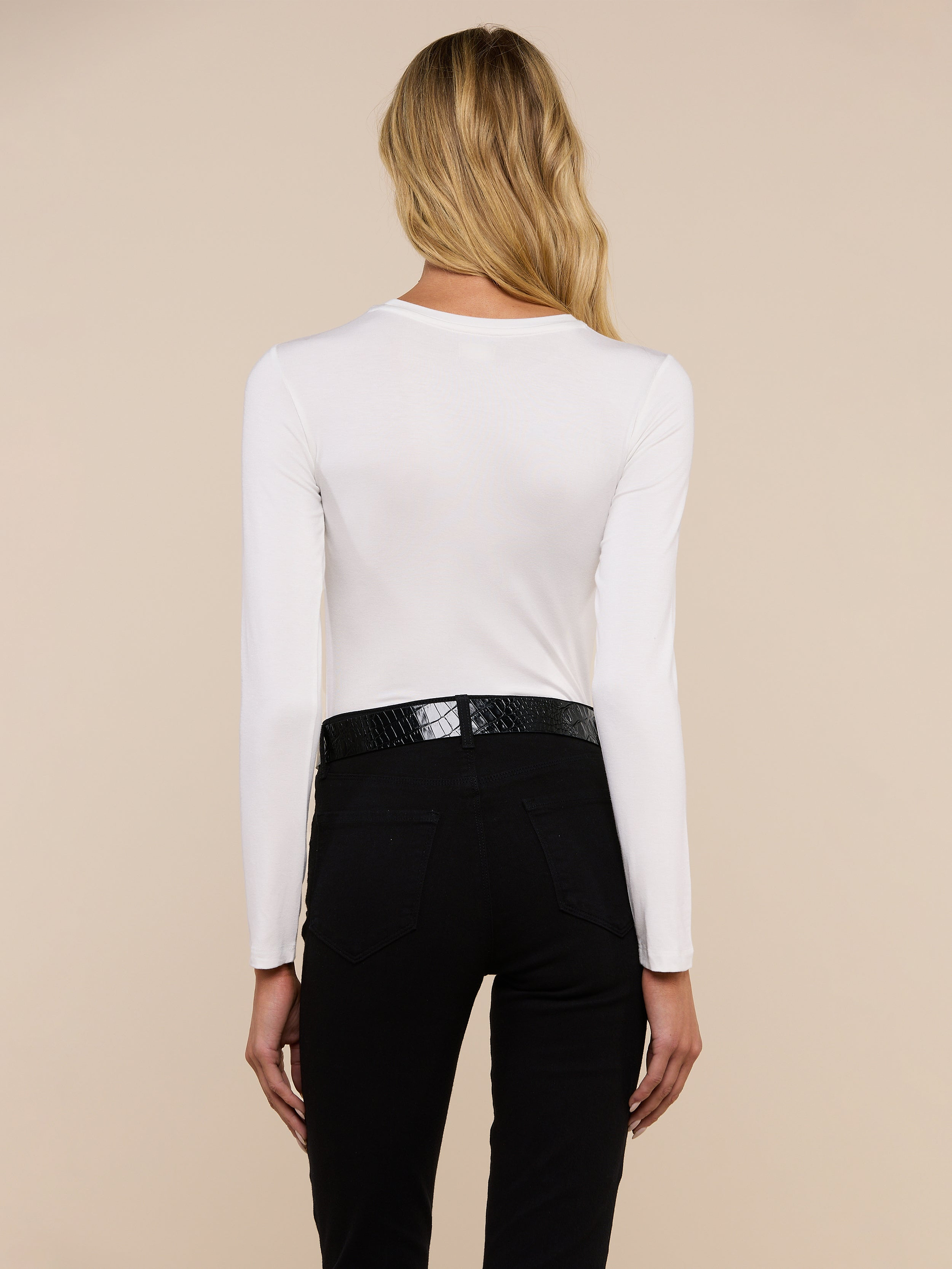 A person with long, wavy blond hair is shown from the back, wearing the Tess Long Sleeve Tee, a black belt, and black pants against a plain light background.