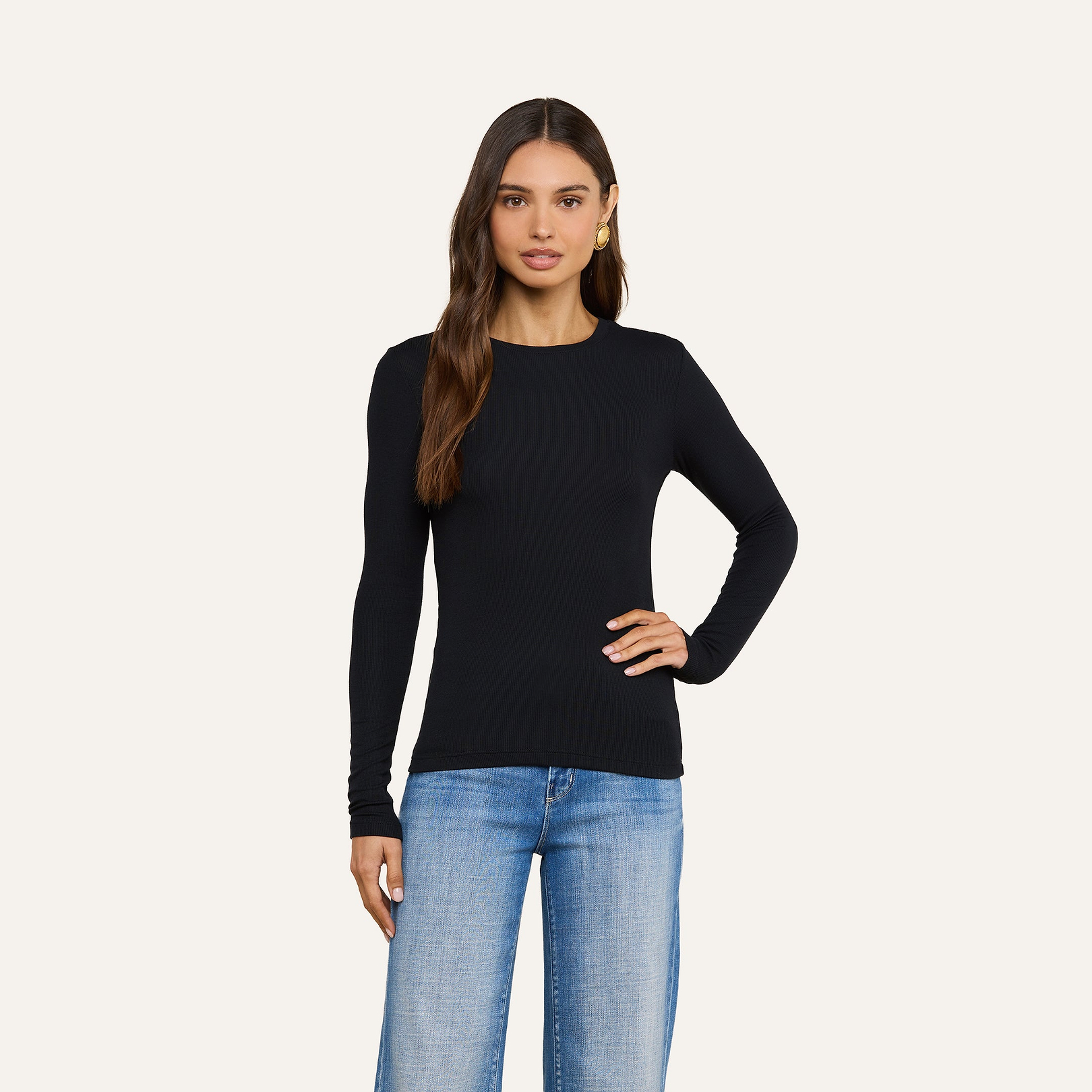 A woman with long brown hair wears the Tess Long Sleeve Tee in black and light blue jeans, standing against a plain beige background with one hand on her hip—a perfect layering piece for any casual look.