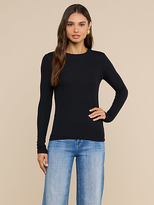 A woman with long brown hair wears the Tess Long Sleeve Tee in black and light blue jeans, standing against a plain beige background with one hand on her hip—a perfect layering piece for any casual look.