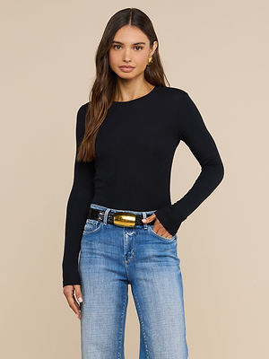 A woman with long brown hair wears the Tess Long Sleeve Tee in black, styled with blue jeans and a black belt with a gold buckle, standing against a plain beige background with one hand in her pocket.