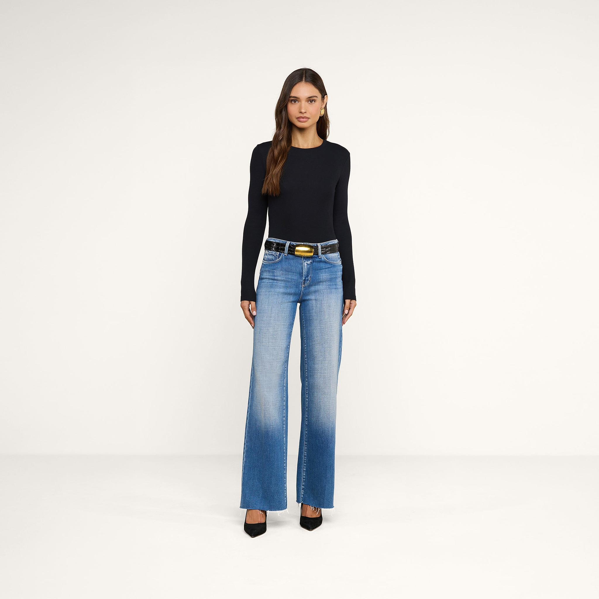 Una donna è in posa su uno sfondo beige e indossa la maglietta a maniche lunghe Tess in nero, abbinata a jeans a vita alta e a gamba larga di colore azzurro, una cintura nera e scarpe con tacco a punta nere.
