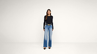 A woman stands in a plain room, wearing a black long-sleeve top, wide-leg blue jeans with a black belt, and black heels.