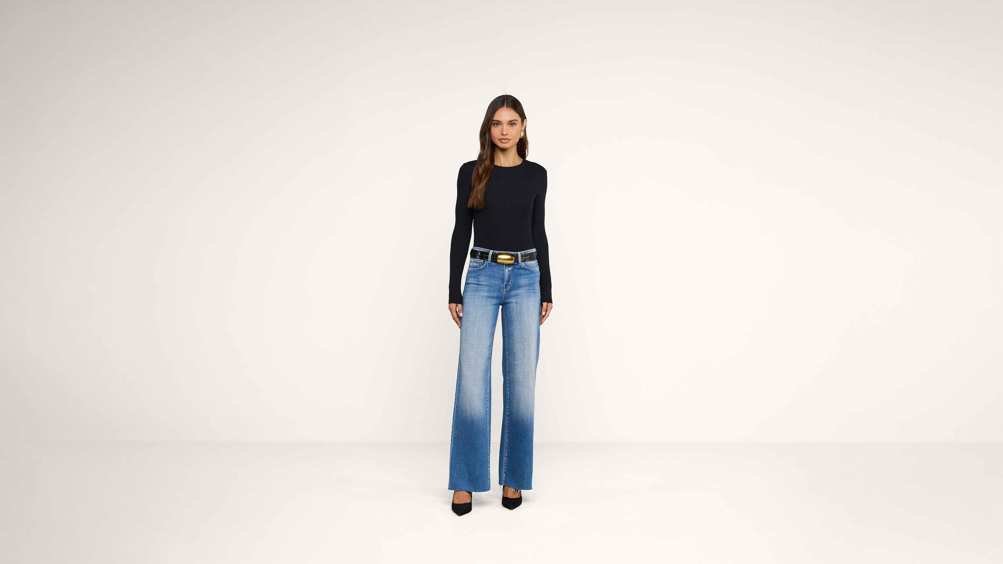 A woman stands in a plain room, wearing a black long-sleeve top, wide-leg blue jeans with a black belt, and black heels.
