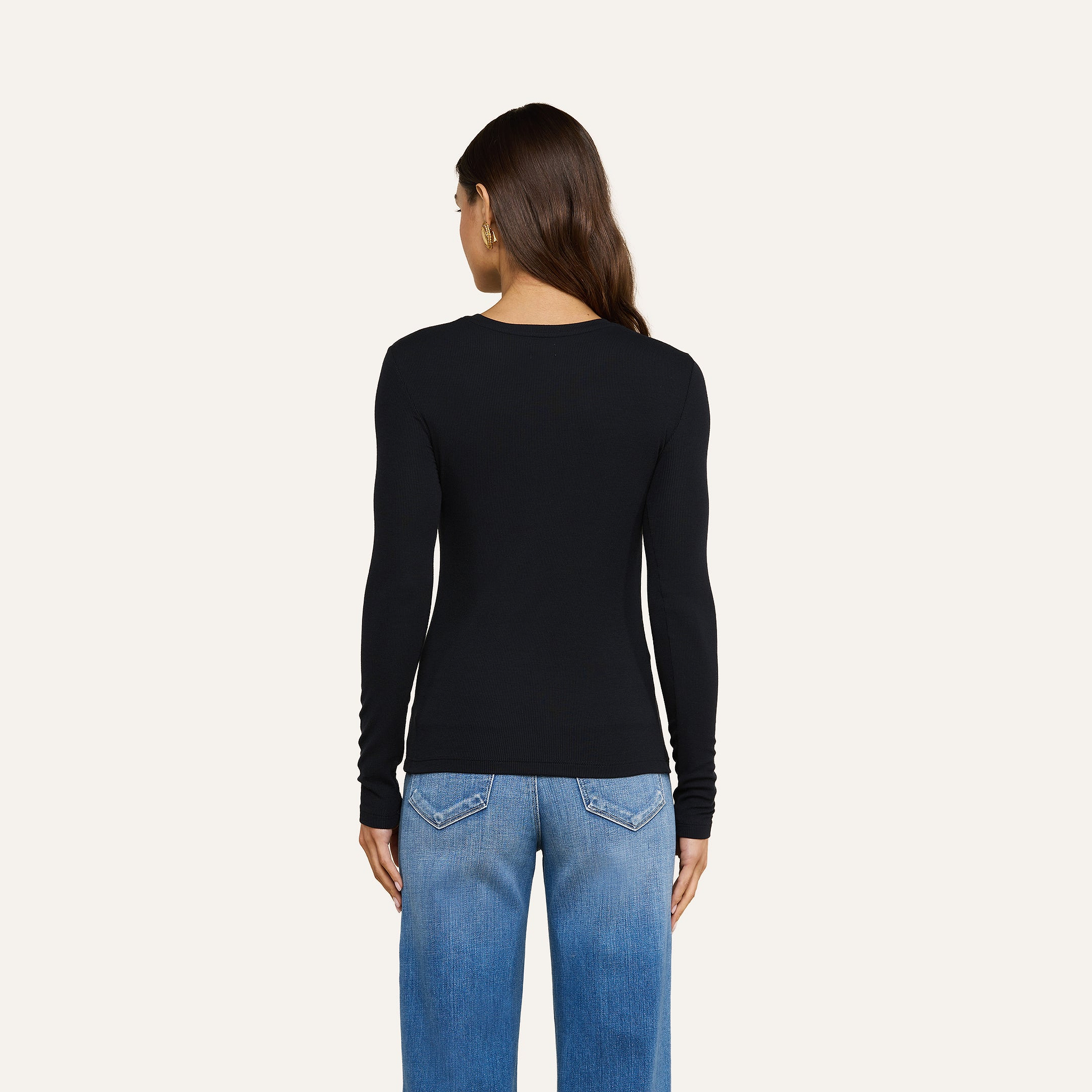 Una donna dai lunghi capelli castani è in piedi di spalle alla fotocamera, indossa la maglietta a maniche lunghe Tess nei colori nero e blu e un paio di jeans, su uno sfondo beige a tinta unita.
