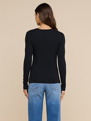 Une femme aux longs cheveux châtains se tient dos à la caméra, vêtue dun t-shirt à manches longues « Tess » noir et dun jean bleu, devant un fond beige uni.