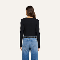 Una donna dai lunghi capelli castani indossa la maglietta a maniche lunghe Tess di colore nero, abbinata a jeans azzurri e una cintura nera, in piedi con le spalle rivolte verso uno sfondo beige.
