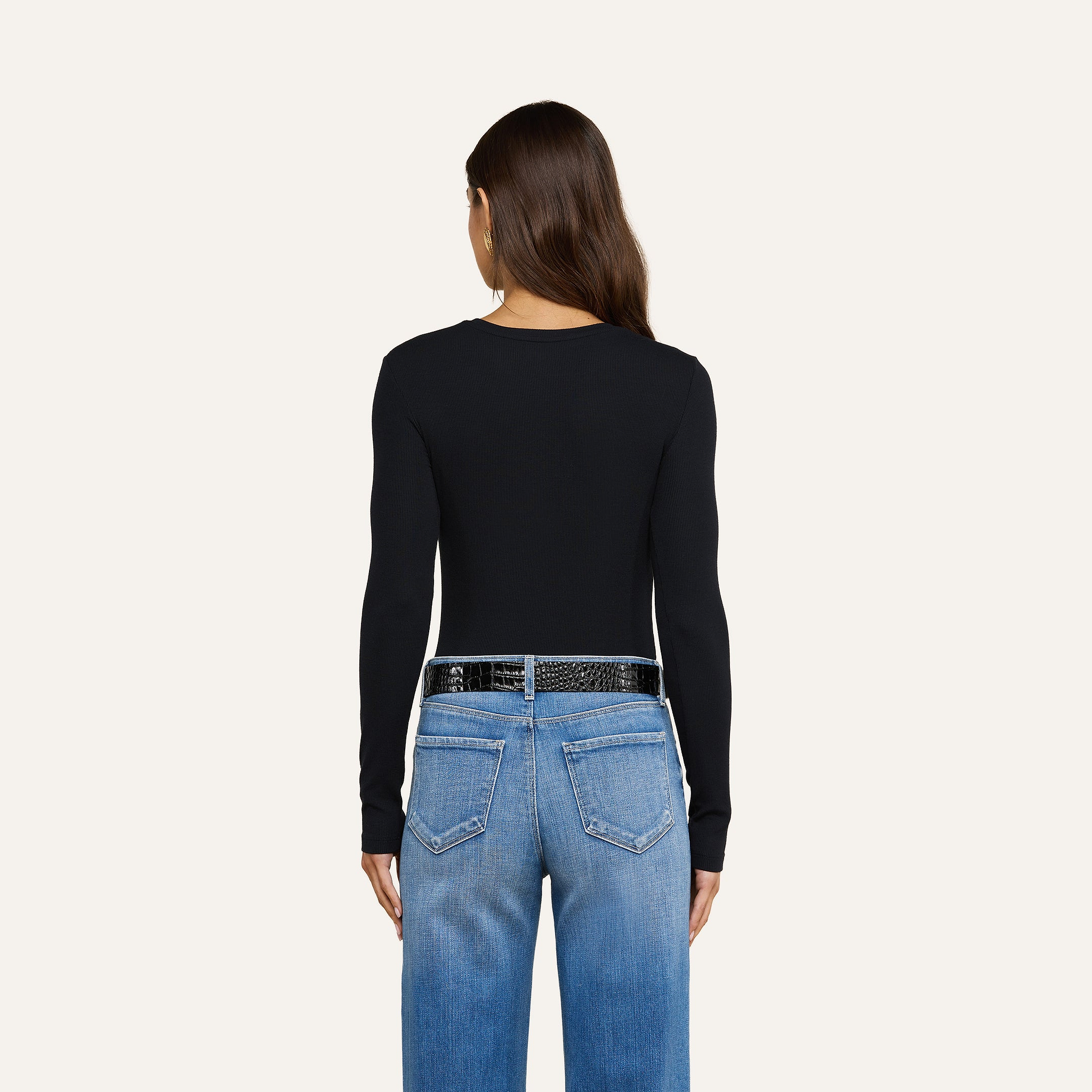Una donna dai lunghi capelli castani indossa la maglietta a maniche lunghe Tess di colore nero, abbinata a jeans azzurri e una cintura nera, in piedi con le spalle rivolte verso uno sfondo beige.