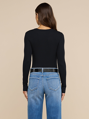 Una donna dai lunghi capelli castani indossa la maglietta a maniche lunghe Tess di colore nero, abbinata a jeans azzurri e una cintura nera, in piedi con le spalle rivolte verso uno sfondo beige.