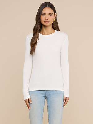 Une femme aux longs cheveux châtains porte le t-shirt à manches longues « Tess » en blanc et un jean bleu clair ; elle se tient debout devant un fond beige, le regard droit devant elle, lexpression neutre.