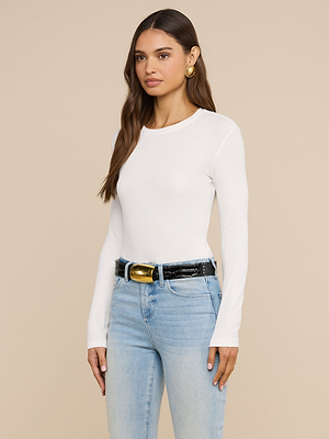 Une femme aux longs cheveux châtains porte un t-shirt à manches longues « Tess » blanc, un jean bleu clair, une ceinture noire à boucle dorée et des boucles doreilles créoles dorées ; elle se tient debout devant un fond beige.