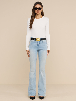Une femme portant des lunettes de soleil, un t-shirt à manches longues Tess blanc, coupe ajustée, un jean évasé bleu clair à taille haute, une ceinture noire à boucle dorée et des talons noirs se tient debout devant un fond beige uni.