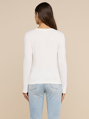 Une femme aux longs cheveux châtains, vêtue dun t-shirt à manches longues « Tess » blanc et dun jean bleu clair, se tient dos à la caméra devant un fond beige.