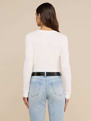 Une femme aux longs cheveux châtains, de dos, porte le t-shirt à manches longues Tess et un jean bleu clair taille haute agrémenté dune ceinture noire, sur un fond beige uni, mettant ainsi en valeur son style moderne et décontracté.