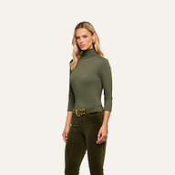 A woman with long blonde hair wears the Aja Turtleneck in soft olive green knit, paired with matching slim fit pants and a wide belt, standing against a plain light background.