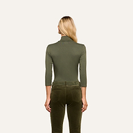 A woman with long blonde hair stands facing away, wearing the Aja Turtleneck in olive green with three-quarter sleeves and slim fit, paired with dark olive velvet pants, against a plain light background.