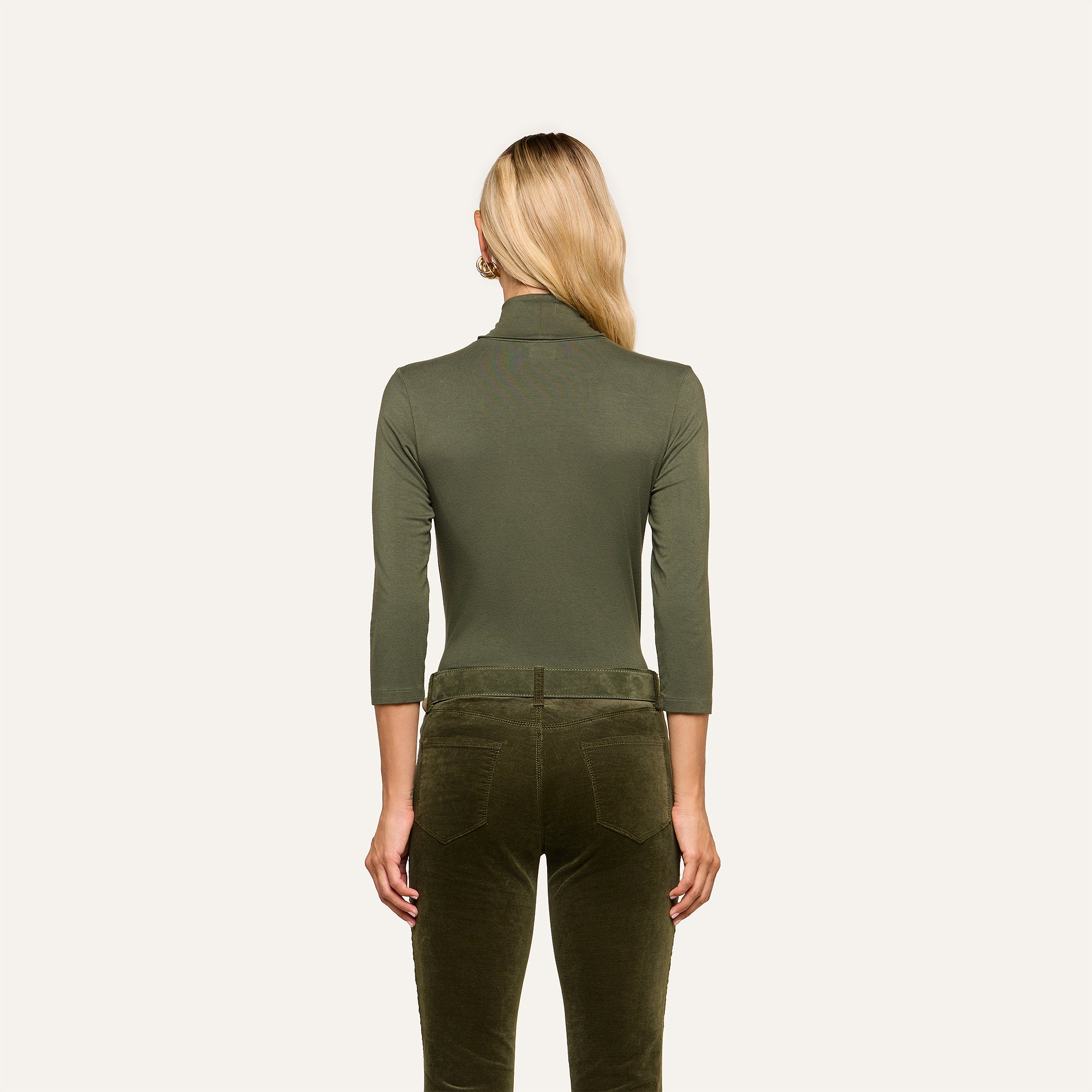 A woman with long blonde hair stands facing away, wearing the Aja Turtleneck in olive green with three-quarter sleeves and slim fit, paired with dark olive velvet pants, against a plain light background.