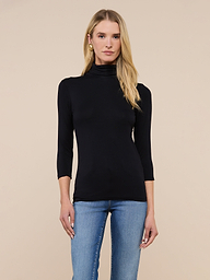 A woman with long blonde hair wears the Aja Turtleneck, a slim-fit black top with three-quarter sleeves, paired with blue jeans and standing against a plain light background.