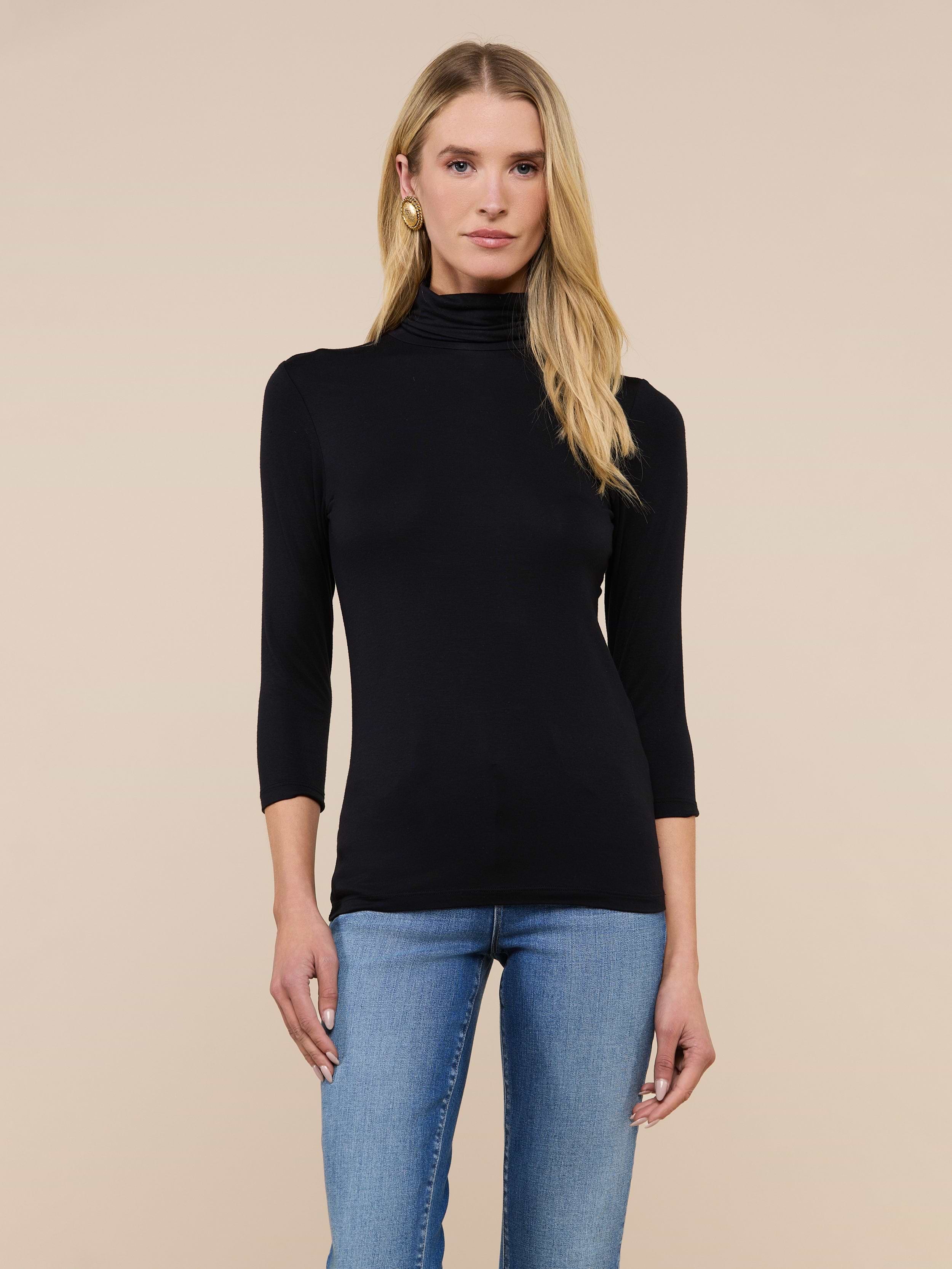A woman with long blonde hair wears the Aja Turtleneck, a slim-fit black top with three-quarter sleeves, paired with blue jeans and standing against a plain light background.