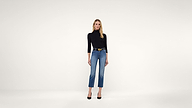 A woman stands against a plain white background, wearing a black long-sleeve top, blue high-waisted jeans, a black belt, and black heels.