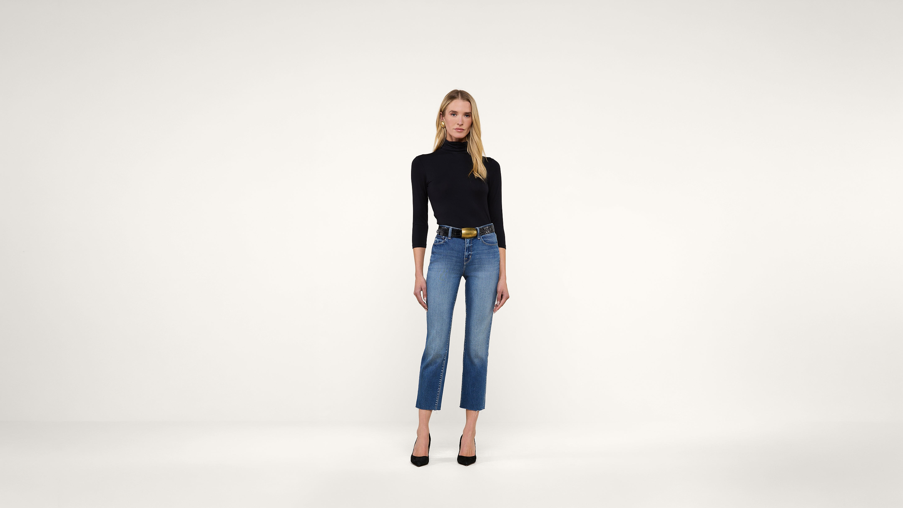 A woman stands against a plain white background, wearing a black long-sleeve top, blue high-waisted jeans, a black belt, and black heels.