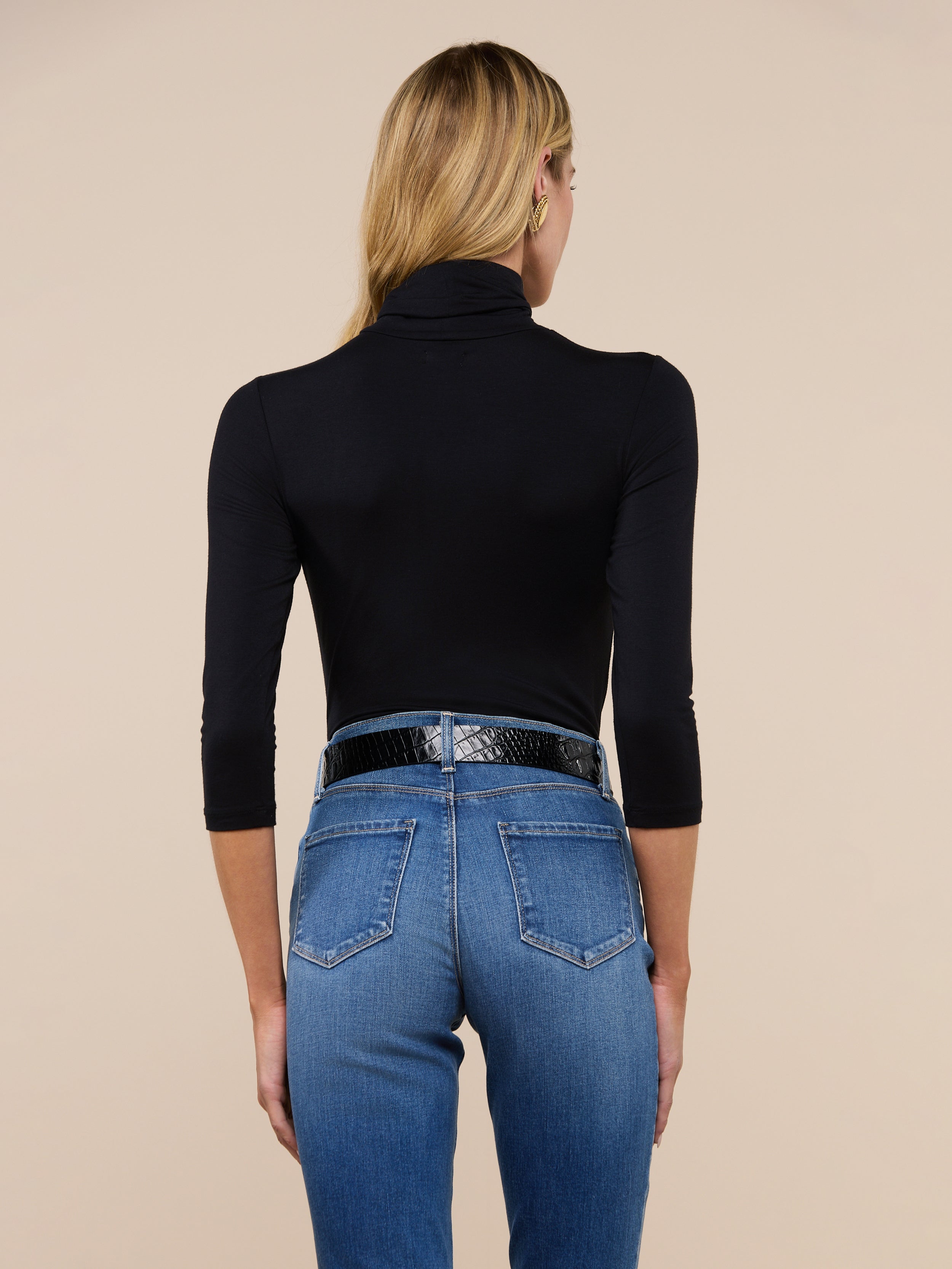 A woman with long blonde hair, wearing the Aja Turtleneck in black along with blue jeans and a black belt, stands with her back to the camera against a plain background.