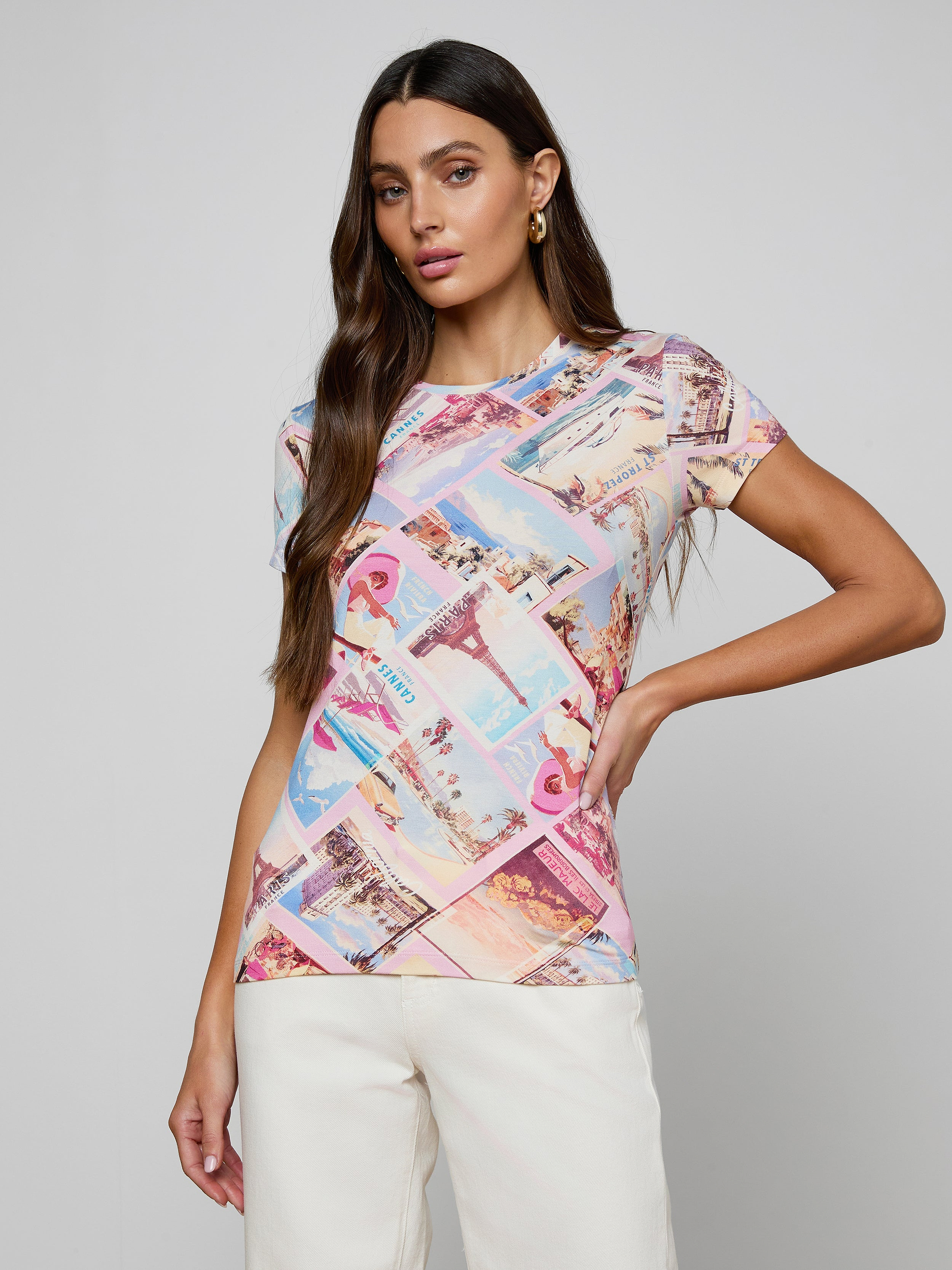 A woman with long brown hair stands confidently against a plain background, wearing the bestselling Ressi Fitted Tee in a colorful postcard print paired with white pants, one hand on her hip as she looks at the camera.