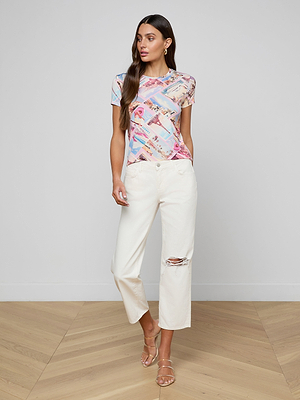 A woman stands indoors wearing the Ressi Fitted Tee in stretch jersey knit, paired with white ripped jeans and strappy sandals. She has long brown hair, her left hand in her pocket as she looks to the side.