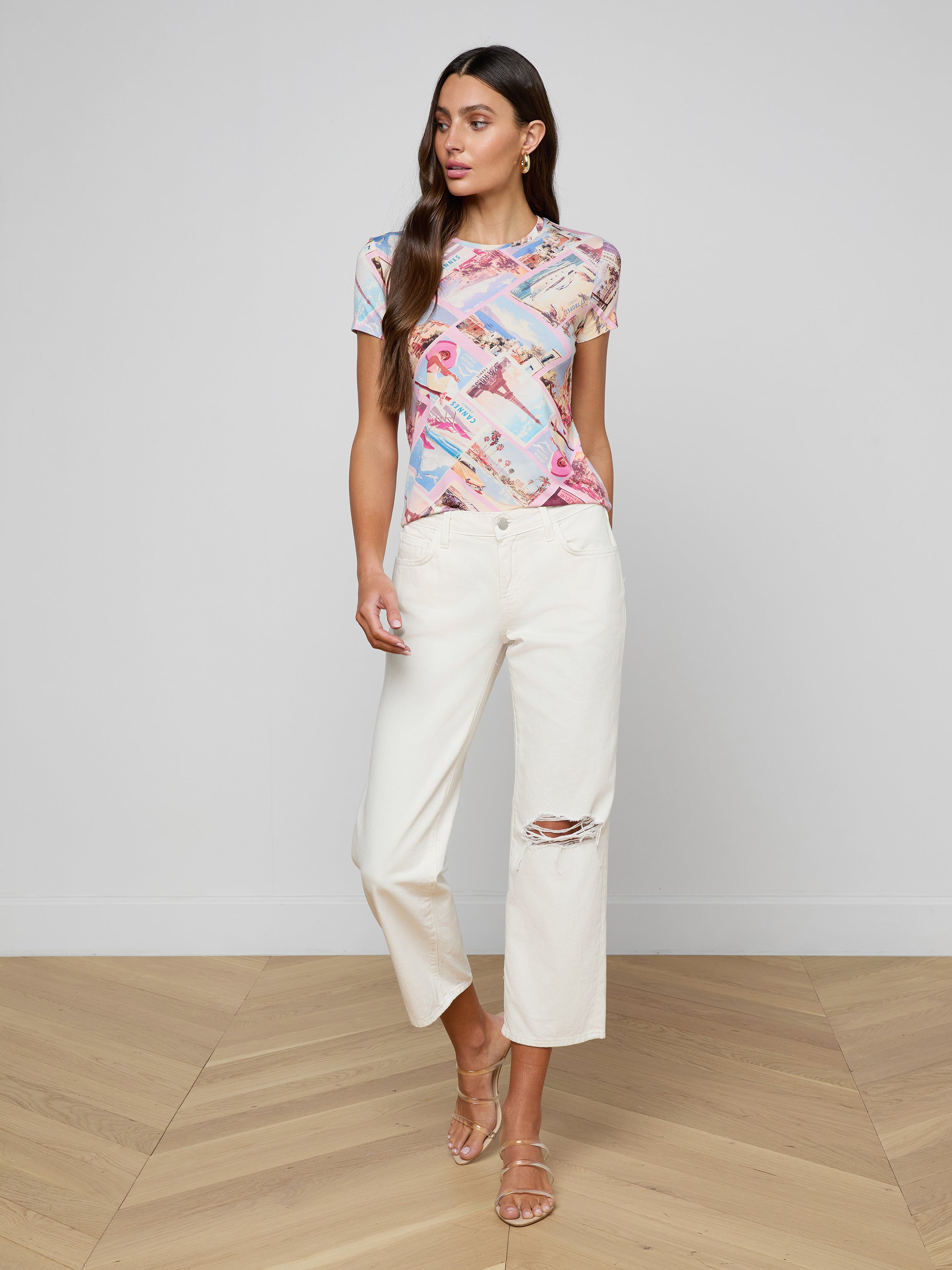Wearing the bestselling Ressi Fitted Tee in a vibrant print, white wide-leg pants featuring a ripped knee, and beige strappy sandals, the woman stands against a plain white background.