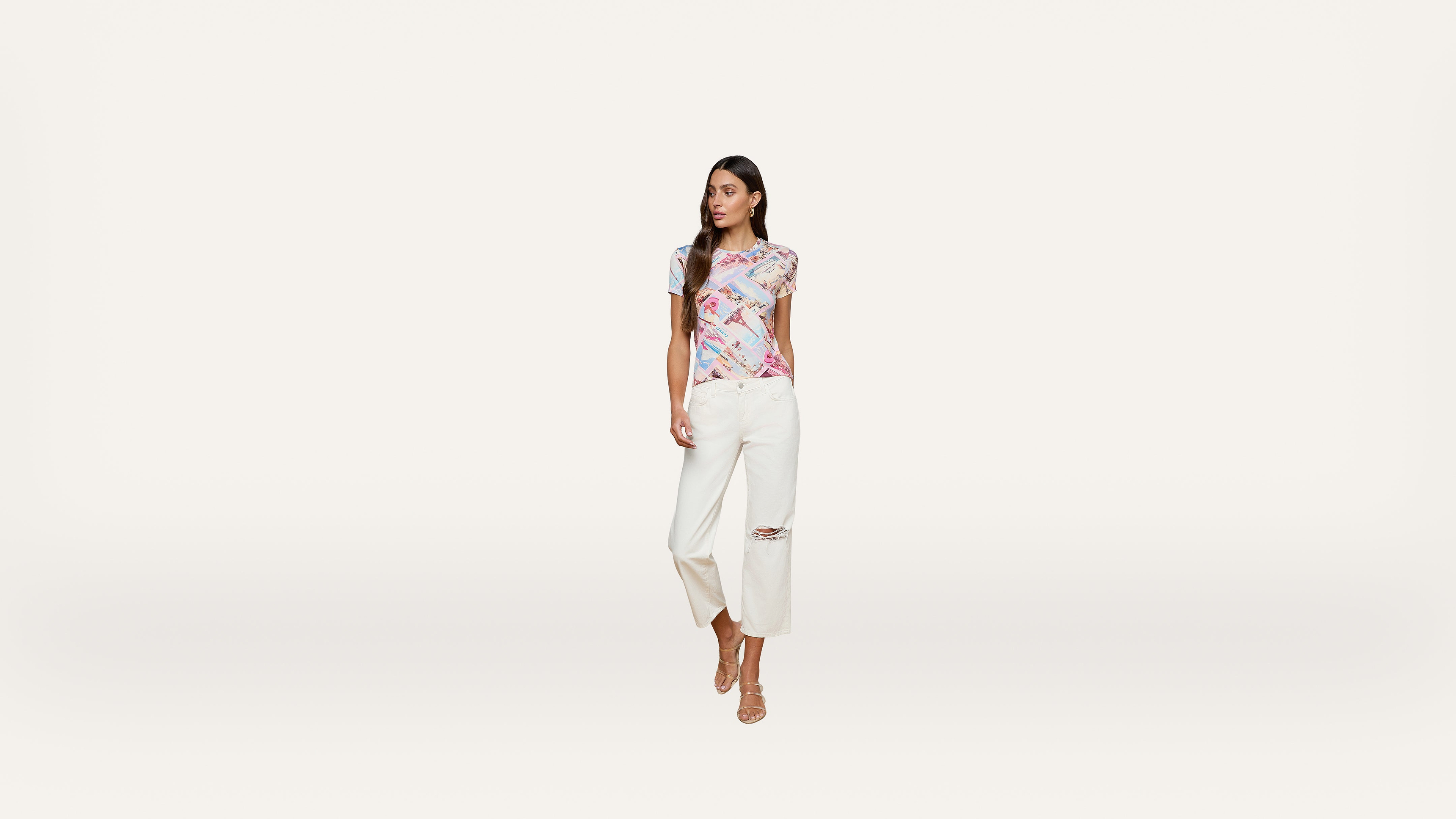 A woman stands against a plain white background, wearing a colorful printed top, white wide-leg ripped jeans, and beige open-toe heels. She looks to the side and has long, straight brown hair.
