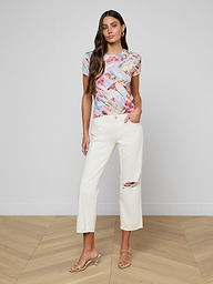 A woman stands against a white background, wearing the bestselling Ressi Fitted Tee in stretch jersey knit, paired with white cropped pants featuring a rip at one knee and beige heeled sandals.