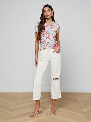 A woman with long brown hair and one hand in her pocket stands against a plain wall wearing the bestselling Ressi Fitted Tee in colorful graphic stretch jersey, slim-cut white high-waisted jeans with a knee rip, and clear heeled sandals.