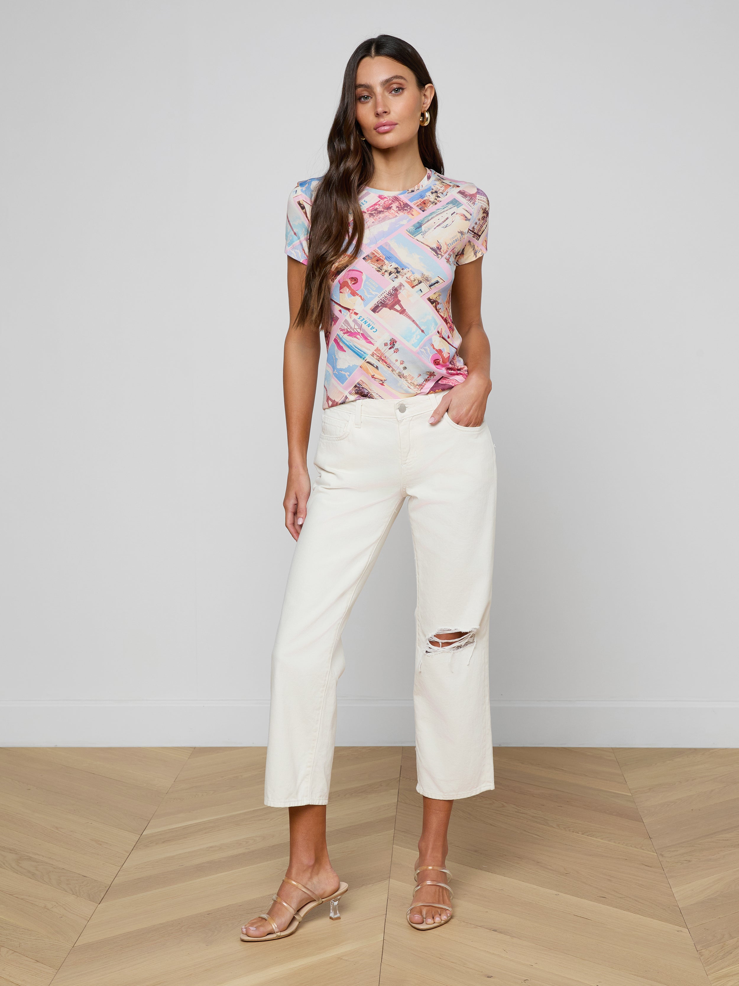A woman stands against a white background, wearing the bestselling Ressi Fitted Tee in stretch jersey knit, paired with white cropped pants featuring a rip at one knee and beige heeled sandals.