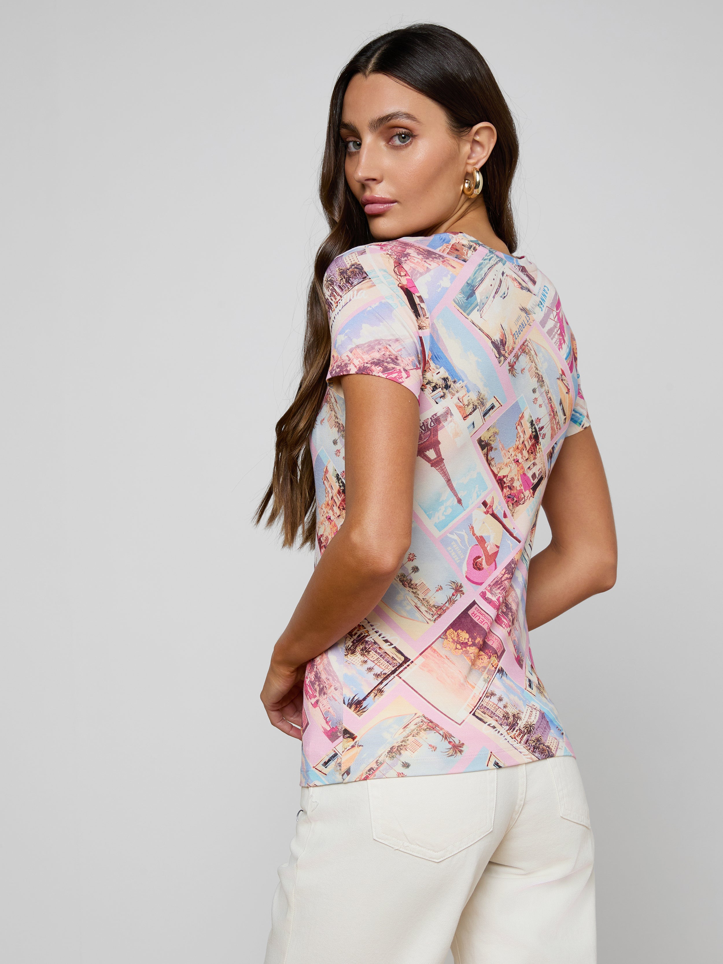 A woman with long brown hair poses with her back turned, looking over her shoulder against a plain, light background. She is wearing the bestselling Ressi Fitted Tee—a colorful short-sleeve top in stretch jersey knit—paired with white pants.