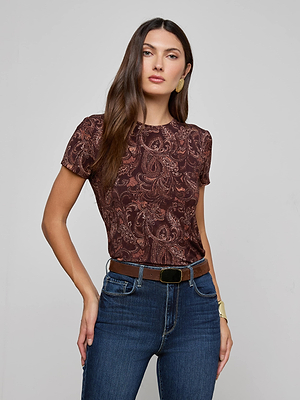 A woman wears the L'AGENCE Ressi Fitted Tee—brown paisley print, stretch jersey knit, short sleeves—with blue high-waisted jeans, brown belt, gold bracelet, and earrings, standing against a plain light gray background.
