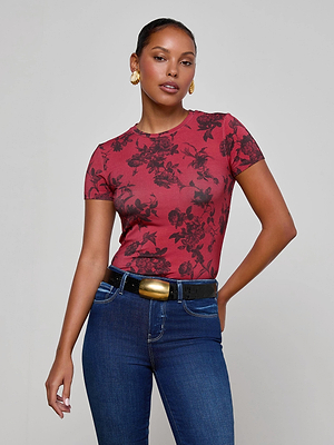 A woman stands confidently against a plain background, wearing the L'AGENCE Ressi Fitted Tee in a burgundy floral print, paired with dark jeans, a black belt with a gold buckle, gold hoop earrings, and her hair pulled back.