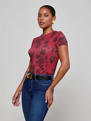 A woman stands against a plain gray background wearing the L'AGENCE Ressi Fitted Tee in burgundy floral print, blue jeans, a black belt with a gold buckle, and gold hoop earrings.