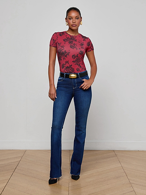 A woman stands in a minimal room wearing the L'AGENCE Ressi Fitted Tee in burgundy floral print, blue bootcut jeans with a wide belt, black pointed shoes, and gold earrings. She faces forward with a neutral expression and relaxed posture.