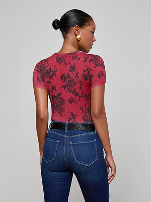 A woman with her hair in a bun, wearing gold hoop earrings, a L'AGENCE Ressi Fitted Tee in burgundy floral print, a black textured belt, and dark blue jeans stands facing away against a plain light gray background.