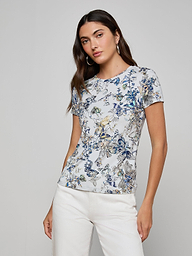 A woman with long dark hair models the Ressi Fitted Tee in a light floral print, paired with white pants. She stands in a relaxed pose against a plain gray background, one hand on her hip.