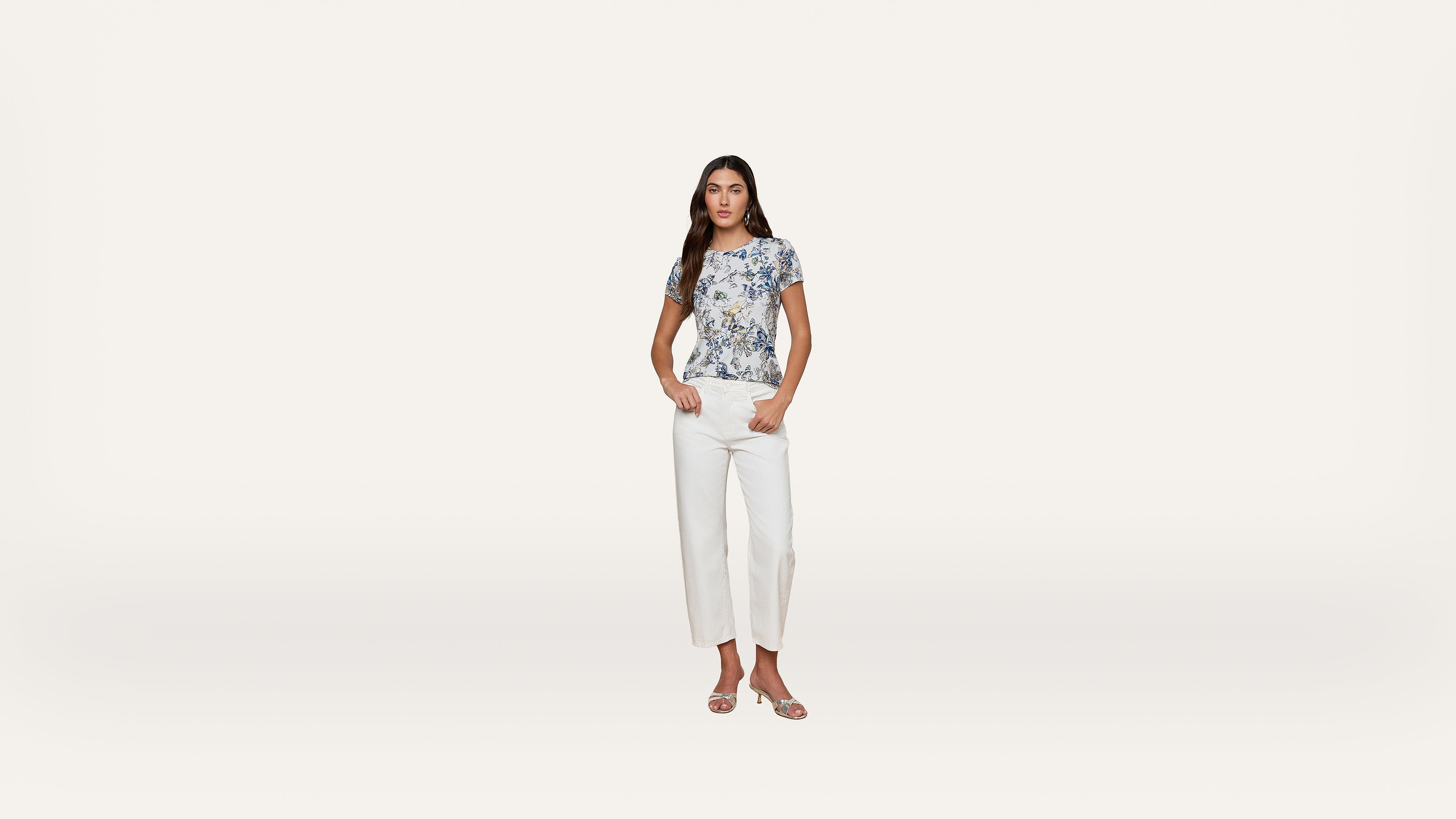 Woman standing against a plain white background, wearing a blue and white patterned short-sleeve top, white pants, and open-toe sandals, with her hands in her pockets.