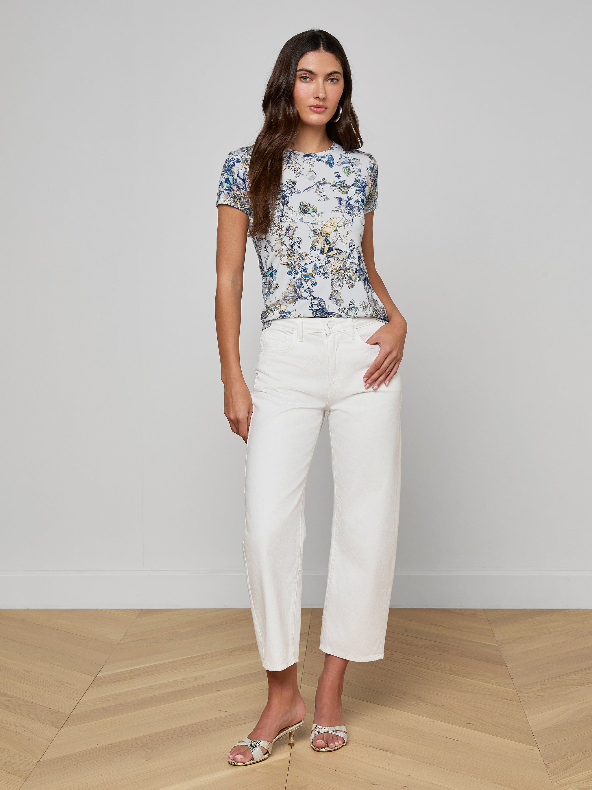 A woman with long brown hair stands indoors on a wooden floor in a relaxed pose against a light gray wall, wearing the Ressi Fitted Tee with a butterfly print, white wide-leg pants, and silver sandals.