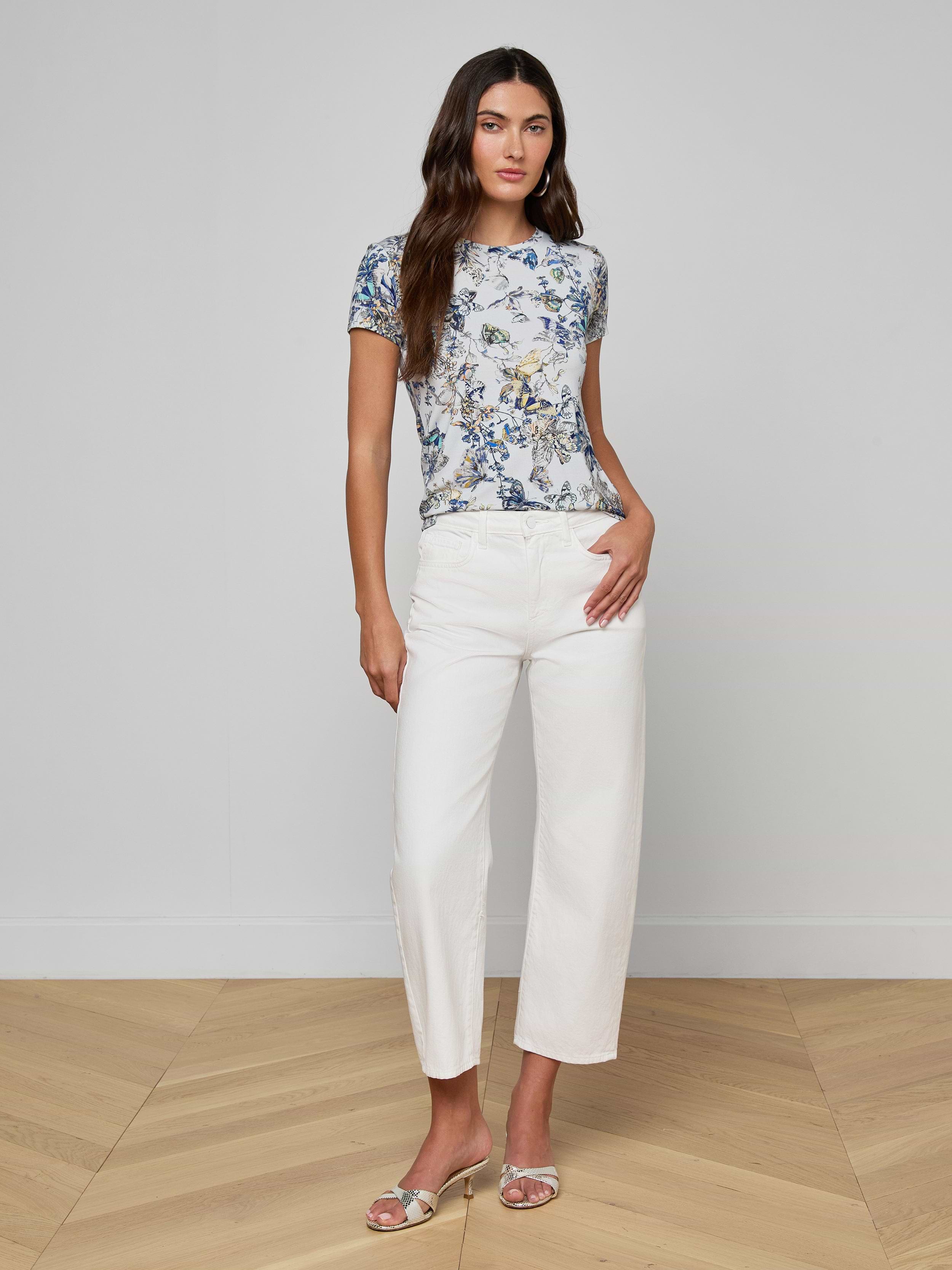 A woman with long brown hair stands indoors on a wooden floor in a relaxed pose against a light gray wall, wearing the Ressi Fitted Tee with a butterfly print, white wide-leg pants, and silver sandals.